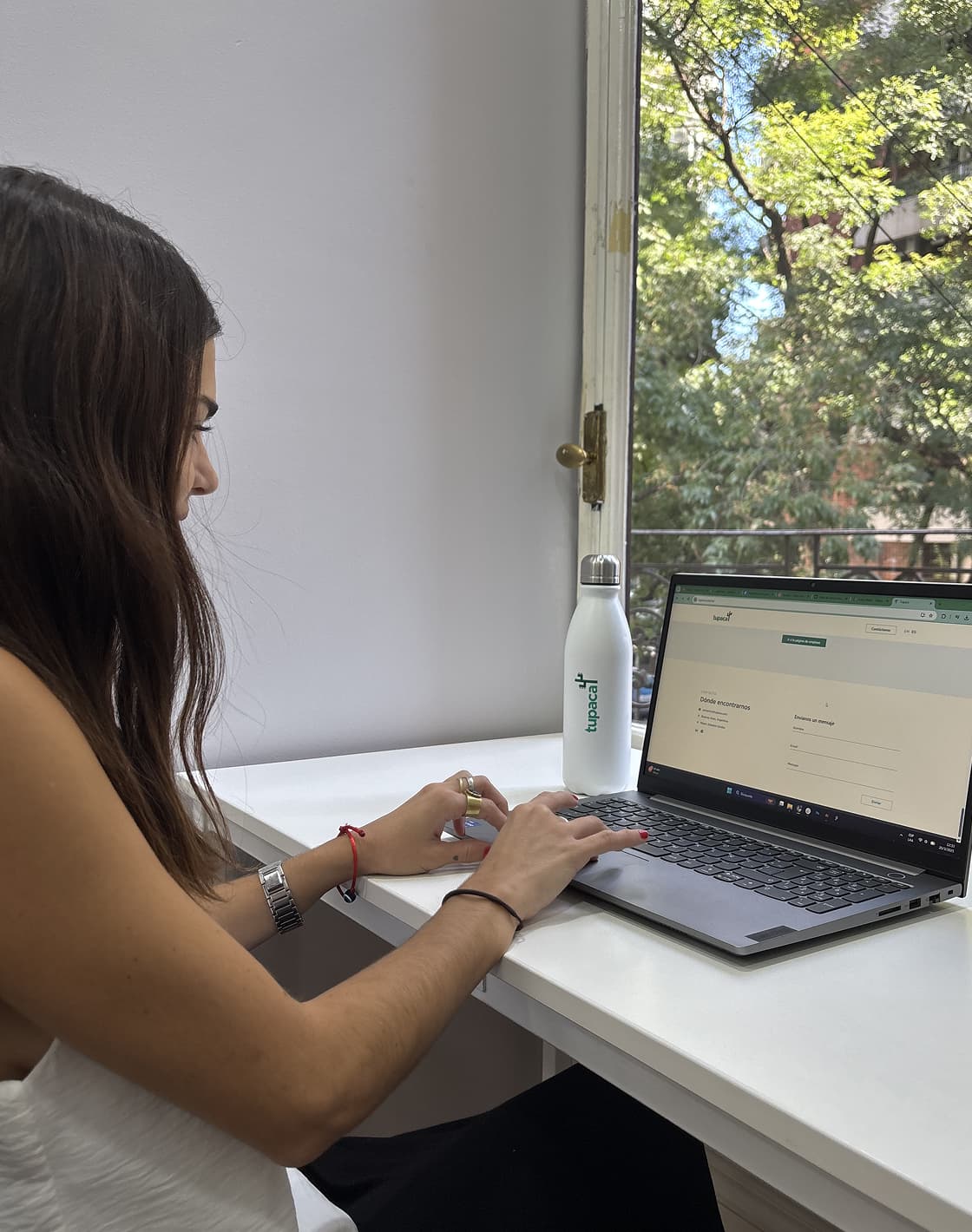Mujer trabajando en un portátil en un escritorio junto a una ventana con vista a árboles